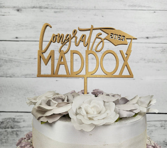 custom wooden cake topper for graduation. cake topper says congrats on top with grad cap & current graduation year, below the cursive congrats, is the graduate's name in all capital letters