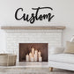May include: A white brick fireplace with a wooden mantel above it. The fireplace has a black metal surround and a wood-burning fire inside. There are several lit candles in front of the fireplace. Above the mantel is a black metal sign that says 'Custom' in a cursive font. A white couch with a tan throw pillow is in the foreground.