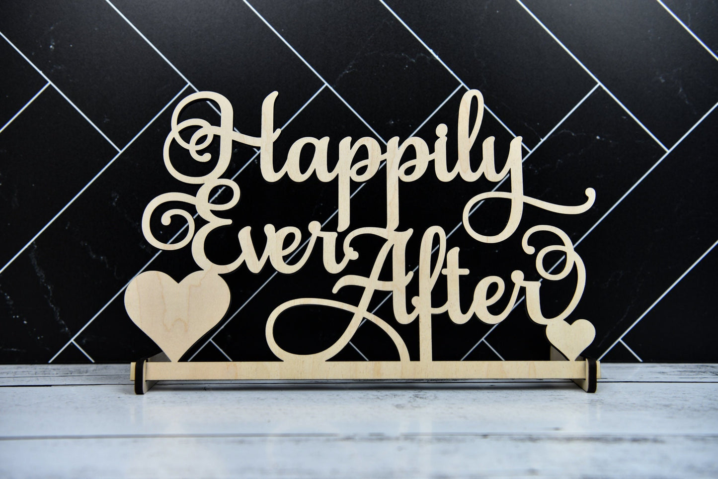May include: Wooden sign with the text 'Happily Ever After' in cursive script. The sign has two small hearts on either side of the text. The sign is on a white wooden surface.