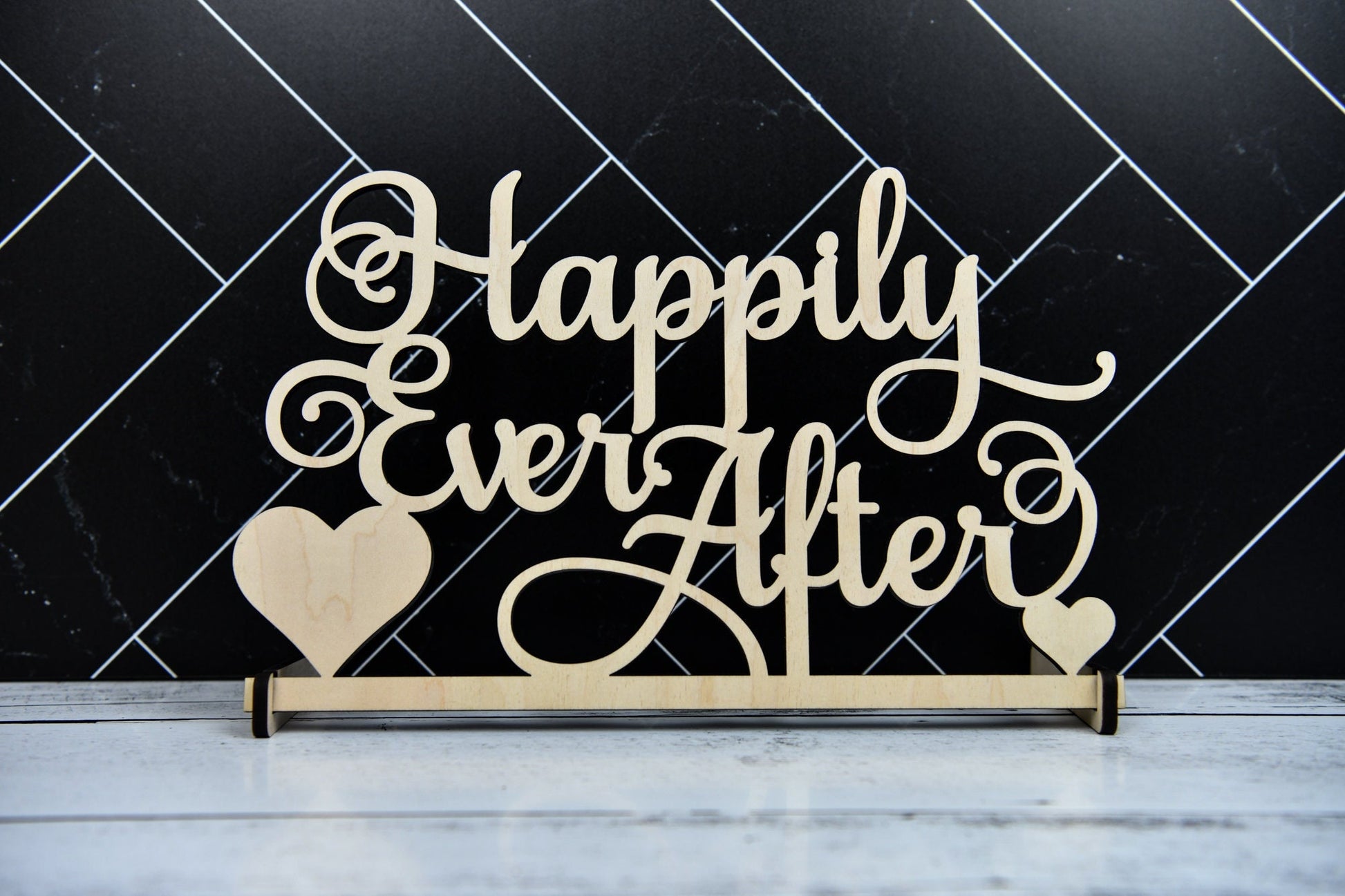 May include: Wooden sign with the text 'Happily Ever After' in cursive script. The sign has two small hearts on either side of the text. The sign is on a white wooden surface.