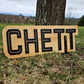May include: A wooden sign with the name "CHETT" in black, stylized lettering. The sign is rectangular with rounded corners and is set against a backdrop of grass and a tree trunk.